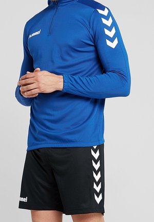 Homme portant un haut de sport bleu à manches longues avec des chevrons blancs sur les manches et un short noir avec des chevrons blancs assortis sur le côté.