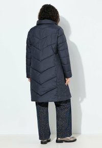 Žena v dlouhém tmavém prošívaném kabátu, vzorovaných širokých kalhotách a kovových botách, otočená zády k jednobarevnému bílému pozadí.