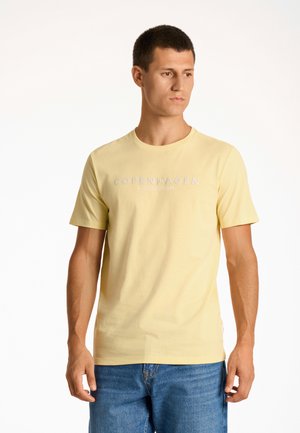 Camiseta de algodón amarilla con cuello redondo, que presenta "COPENHAGUE LINDGBRGH" impreso en blanco en el pecho. Combinada con pantalones cortos de mezclilla azul.