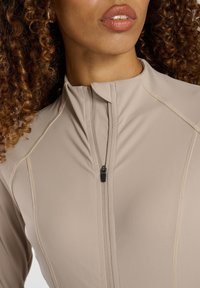 Jasno beżowa bluzka zapinana na zamek, wykonana z gładkiego materiału, z wysokim kołnierzem, płaskimi szwami oraz małym zamkiem przy szyi. Widoczne kręcone włosy.