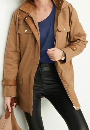 Femme portant une veste à capuche beige sur une chemise bleue et un pantalon en cuir noir, tenant un sac beige avec des poignées en cuir marron.
