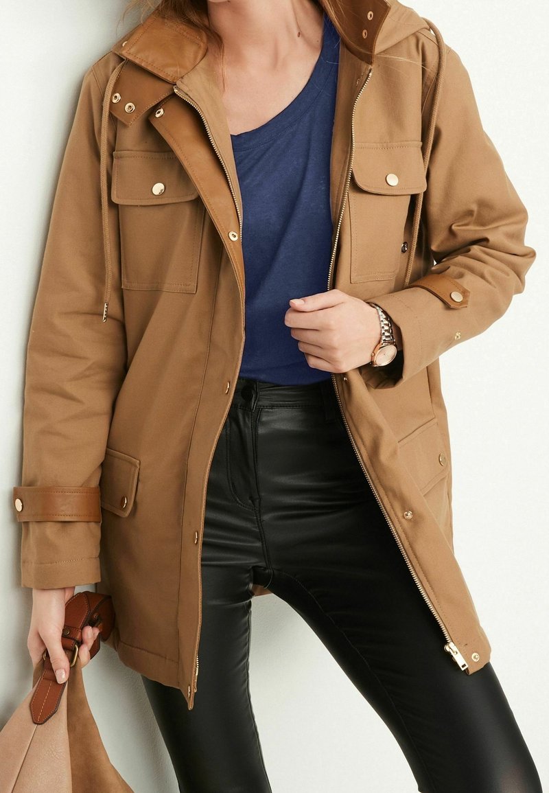 Femme portant une veste à capuche beige sur une chemise bleue et un pantalon en cuir noir, tenant un sac beige avec des poignées en cuir marron.