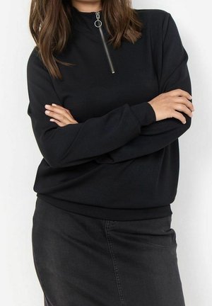 Femme portant un haut noir à manches longues avec une fermeture éclair à l'avant et une jupe noire, les bras croisés, devant un fond blanc uni.