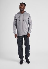 Grijze rits-hoodie met een capuchon, zwart logo op de borst, gecombineerd met zwarte, lichte sportbroek en zwarte sneakers.