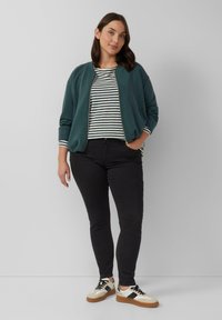Donkergroene bomberjack over een gestreept shirt, zwarte skinny jeans en witte sneakers met tan accenten. Zachte texturen, casual ontwerp.