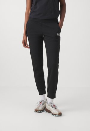 Persoon draagt zwarte EA7 joggingbroek, witte sokken en beige en bruine sneakers, staand met één hand in de zak tegen een effen achtergrond.