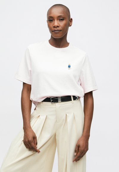 Osoba s krátkými vlasy, oblečená v světlém růžovém tričku s malým vyšitým foxem a krémových plisovaných kalhotách s černým páskem.