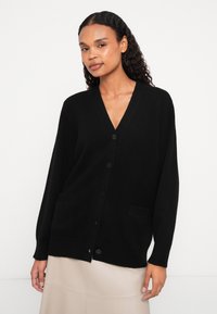 Cardigã preto com decote em V, mangas longas, bolsos frontais e fecho de botão; feito de um material suave e texturizado.