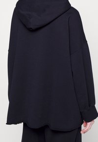 Felpa nera oversize realizzata in un tessuto morbido e ricco. Presenta una vestibilità rilassata, maniche lunghe e un orlo texturizzato. Cappuccio sul retro.