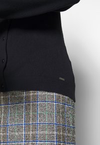 Schwarzer Strickpullover mit geripptem Saum und Knopfdetails, kombiniert mit einem karierten Rock in Braun-, Blau- und Grüntönen. Subtiler Logoakzent.