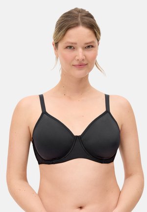 Mujer de piel clara y cabello rubio que lleva un sujetador negro con aro, mirando hacia adelante sobre un fondo blanco liso.