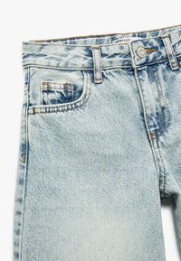 Jeans de mezclilla azul claro con un acabado desgastado, que presentan costuras naranjas contrastantes, un cierre frontal de botón y dos bolsillos laterales.