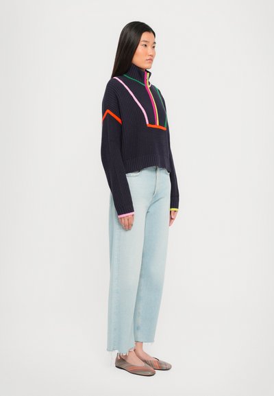 Marineblauwe gebreide pullover met hoge kraag, met kleurrijke accenten in roze, groen en oranje, gecombineerd met lichtblauwe wijde jeans.
