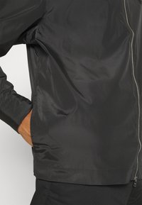 Sort lynlåst jakke med en persons hånd placeret inde i venstre lomme, synlig fra talje til midt brystområde.