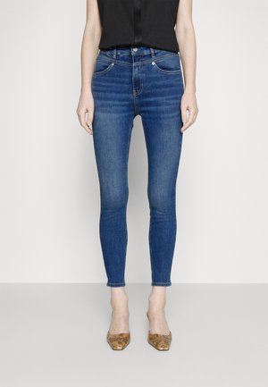 Vrouw draagt blauwe high-waist skinny jeans, zwart mouwloos topje en bruine schoenen met slangenhuidprint en spitse neus, staand tegen een effen achtergrond.