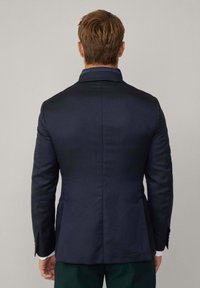 Blazer navy in tessuto testurizzato, vestibilità su misura, colletto con punta, e spacco posteriore singolo, dettagliato con bottoni marroni e polsini della camicia bianchi in vista.