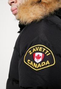 Person, die eine schwarze Jacke mit einer gefütterten Kapuze und einem gelb umrandeten Aufnäher mit der Aufschrift "Zavetti Canada" und einem roten Ahornblatt-Emblem trägt.