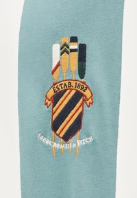 Κεντημένο διακριτικό που απεικονίζει πέντε κουπιά σε διάφορα χρώματα πάνω από μια ασπίδα με διαγώνιες ρίγες, σε φόντο τιρκουάζ ύφασμα.