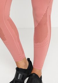 Detail nohou v růžových legínách s vzorovanými síťovými prvky a černými sportovními botami na světlém pozadí.