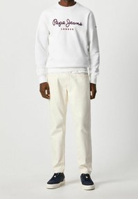 Man i en vit Pepe Jeans London-tröja, krämfärgade byxor och mörkblå sneakers, som står mot en helt vit bakgrund.