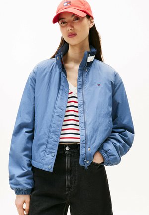 Hellblau bomberjacke mit Reißverschluss und Druckknöpfen, kombiniert mit einem gestreiften V-Ausschnitt-Pullover. Das Model trägt eine rote Mütze. Schwarze Jeans.