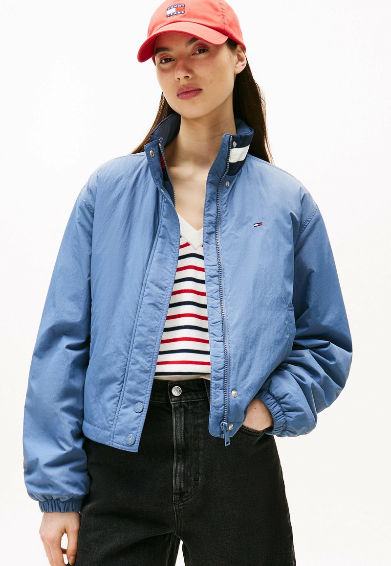 Jasnoniebieska kurtka bomberka z zamkiem błyskawicznym i zatrzaskami, w połączeniu z paskowanym swetrem z dekoltem w serek. Model nosi czerwoną czapkę. Czarne dżinsy.