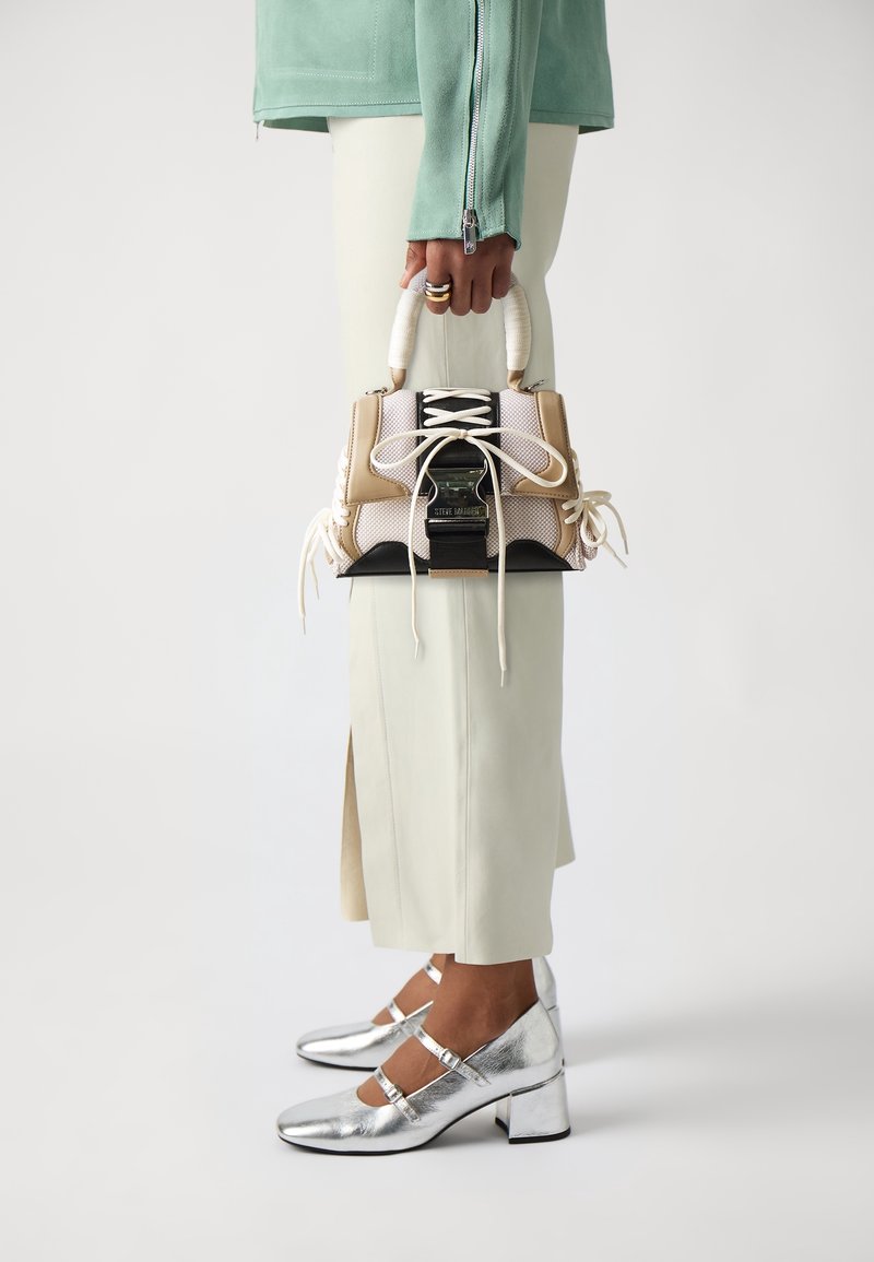 Femme tenant un sac à main beige et blanc avec dentelle, portant des chaussures argentées à petit talon et une longue jupe crème sur fond blanc.