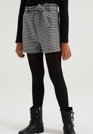 Personne portant un short pied-de-poule noir et blanc avec ceinture à la taille, des collants noirs, un haut noir à manches longues et des bottines noires à boucle, debout sur un fond blanc.