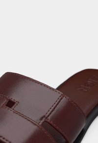 Gros plan sur une lanière de sandale en cuir marron avec des découpes rectangulaires et des coutures visibles sur un fond blanc.