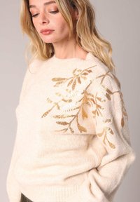 Crème kleurige gebreide trui met een gouden, met pailletten versierd bladerenpatroon op de linker schouder. Zachte textuur, ronde halslijn en losse pasvorm.