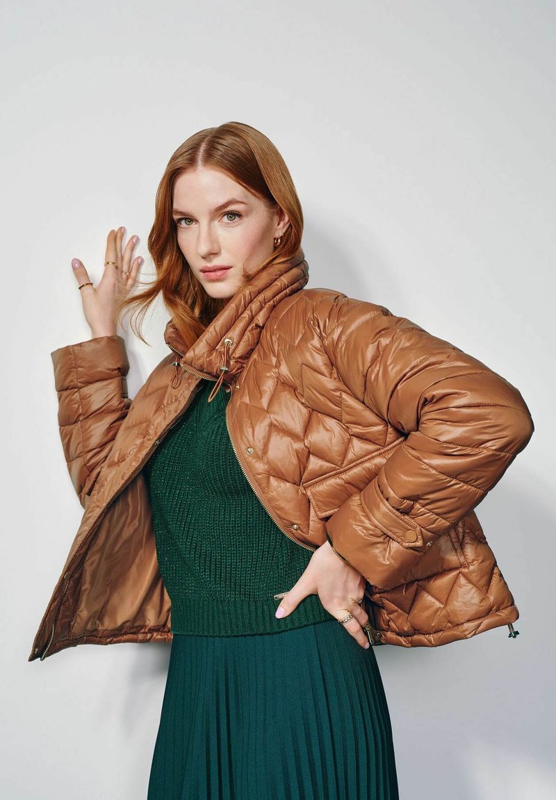 Brązowa pikowana kurtka puchowa z wysokim kołnierzem, zapięciem na zamek i elastycznymi mankietami. Noszona na zielonym swetrze z prążkowanym wzorem i plisowanej spódnicy.