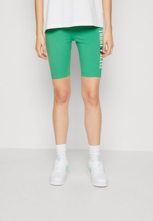 Personne portant un short vert mi-cuisse avec le logo Tommy Jeans sur la jambe gauche, des chaussettes blanches, des baskets blanches et une chemise blanche.