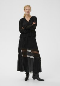 Čierne plisované dlhé šaty s vrstveným dizajnom, obsahujúce priehľadné časti. Dlhé rukávy a voľný strih v páse. Čierne špicaté členkové čižmy.