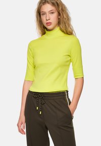 Bright neon yellow short-sleeve turtleneck top; fitted design; smooth texture paired with dark athletic pants featuring stripe accents.