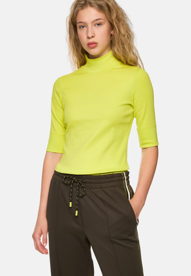 Bright neon yellow short-sleeve turtleneck top; fitted design; smooth texture paired with dark athletic pants featuring stripe accents.