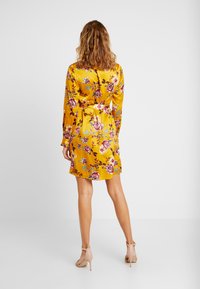 Gul blommig klänning i satinmaterial, med långa ärmar, ett knyt i midjan och ett livligt ros-mönster. Bärs med nude högklackade skor.
