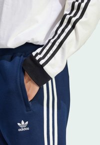 Tumši zili sporta bikses ar baltām trim svītrām gar sāniem. Melni piedurkņu gali un redzama kabata. Adidas logo baltā krāsā.