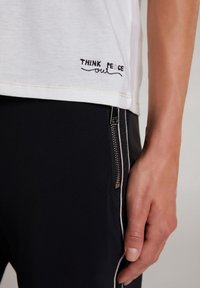 Wit katoenen T-shirt met zwarte geborduurde tekst "THINK PEACE out" aan de zoom, gecombineerd met een zwarte broek met een ritsvak.