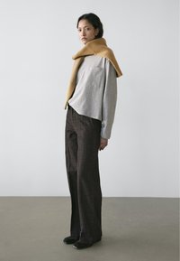 Chemise grise à manches longues avec une poche, associée à un pantalon à carreaux marron et une écharpe moutarde côtelée drapée sur les épaules. Chaussures plates.