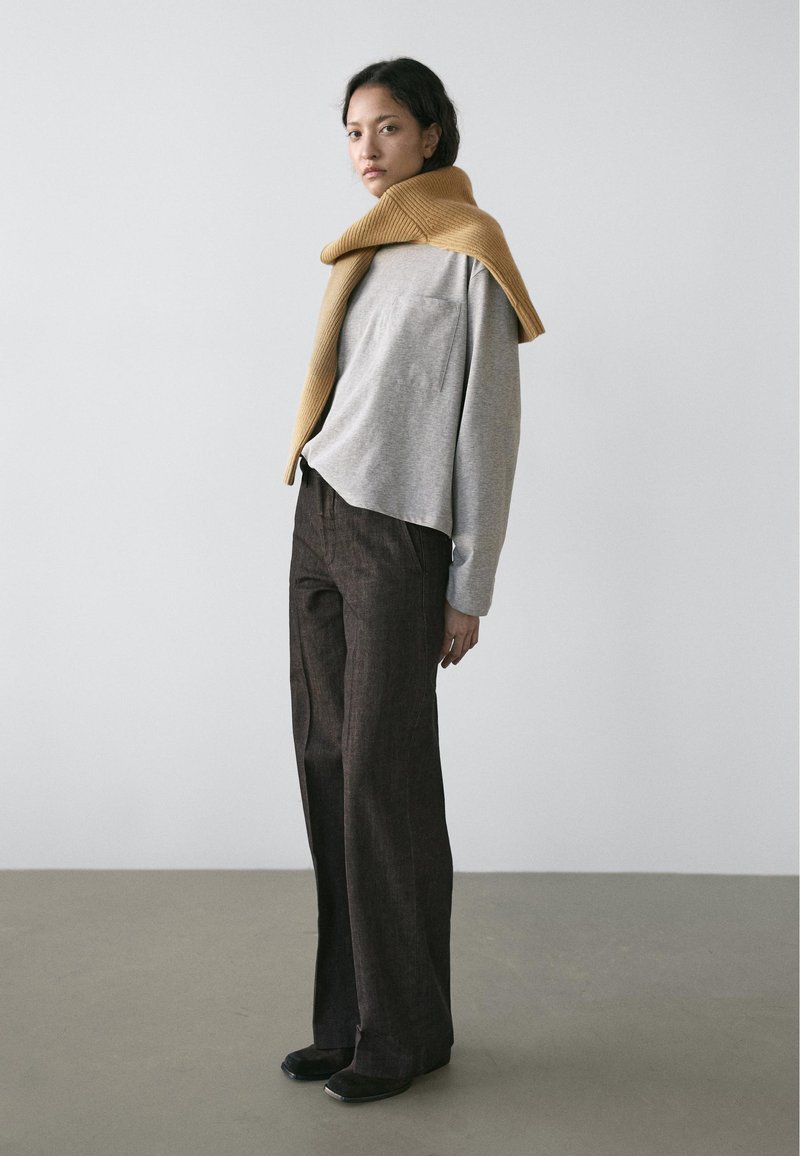 Chemise grise à manches longues avec une poche, associée à un pantalon à carreaux marron et une écharpe moutarde côtelée drapée sur les épaules. Chaussures plates.