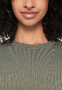 Ελαιοπράσινη ριμπ μανικετό κορυφή με φαρδύ στρογγυλό λαιμό. Η υφή του υφάσματος είναι λεία με κατακόρυφες γραμμές και ελαφριά γυαλάδα.