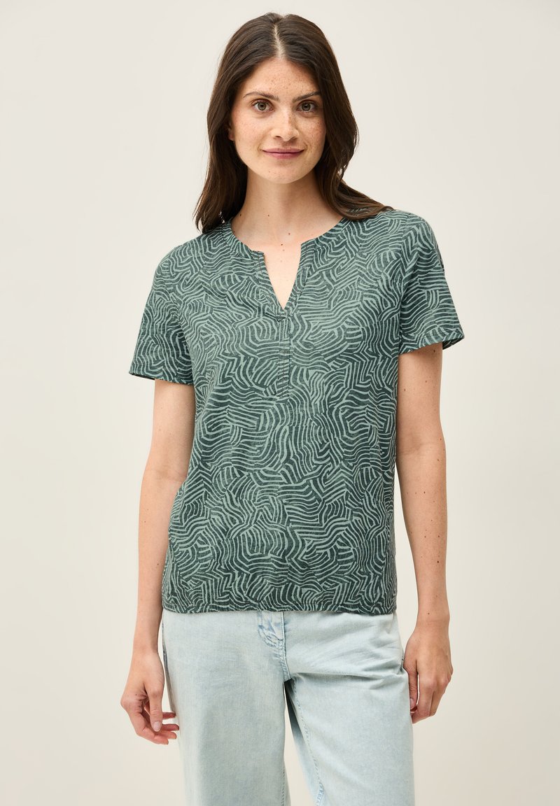 Grünes Kurzarm-Oberteil mit V-Ausschnitt, das ein gewelltes Linienmuster und eine weiche Textur aufweist, kombiniert mit hellblauen, lässig sitzenden Jeans.
