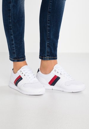 Legs in dark blue jeans wearing white sneakers with red and navy side stripes, standing on a light gray floor.