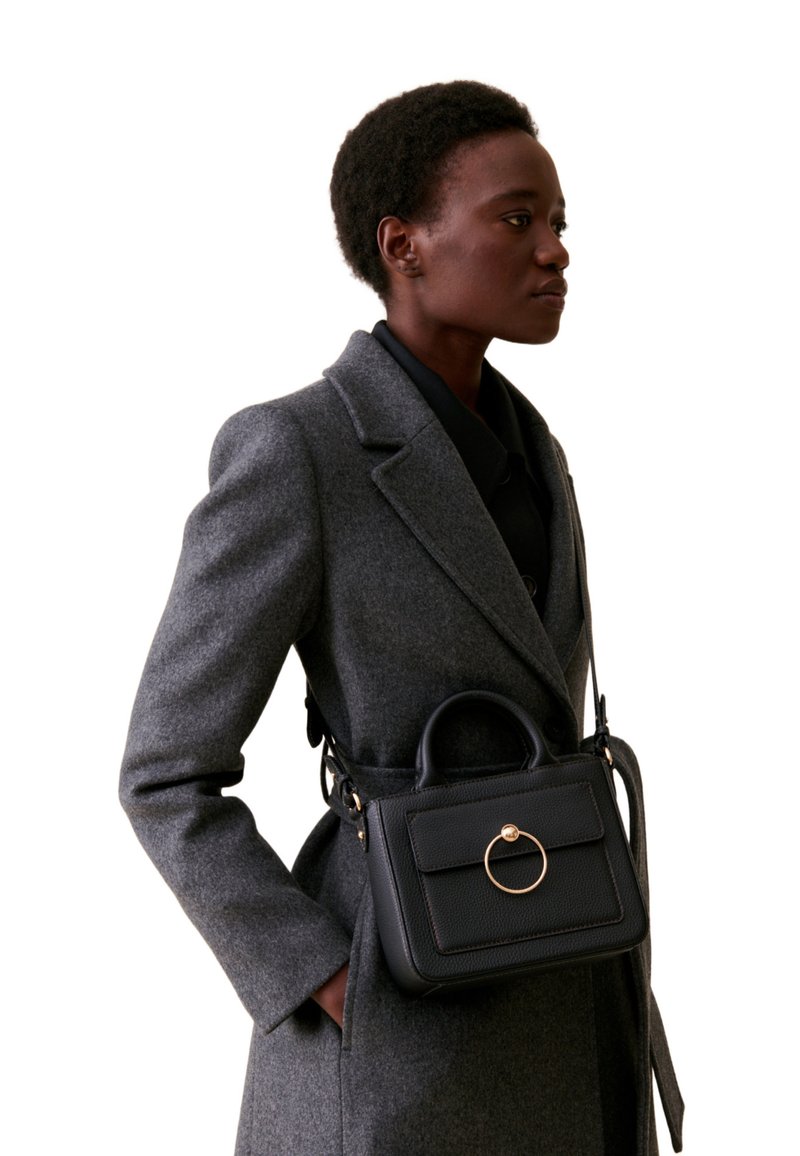 Sac à main en cuir noir avec une poignée supérieure arrondie et des accents en matériel doré. Surface texturée et forme rectangulaire, porté sur un manteau gris.