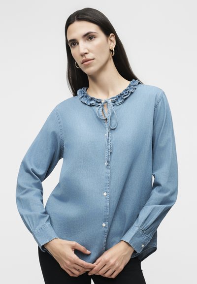 Γυναίκα με μακριά σκούρα μαλλιά που φοράει χρυσούς κρίκους στα σκουλαρίκια και ένα ανοιχτό μπλε πουκάμισο με κουμπιά, γιακά με βολάν και κορδόνι στο λαιμό.