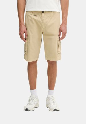 Uomo con pantaloncini cargo beige, calzini bianchi e sneakers bianchi, in piedi con le mani rilassate lungo i fianchi su sfondo uniforme.