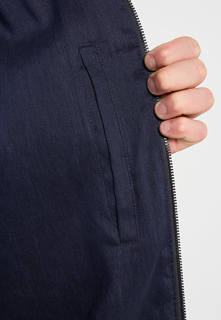 Innenbereich einer marineblauen Jacke mit glatter Textur, Reißverschluss und einer funktionalen Seitentasche. Sichtbare Nähte und Stoffglanz.