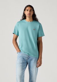 Lyseblå bomulls t-skjorte med korte ermer og rund hals; har en liten rød Levi's-logo på brystet. Brukt med falmete blå jeans.