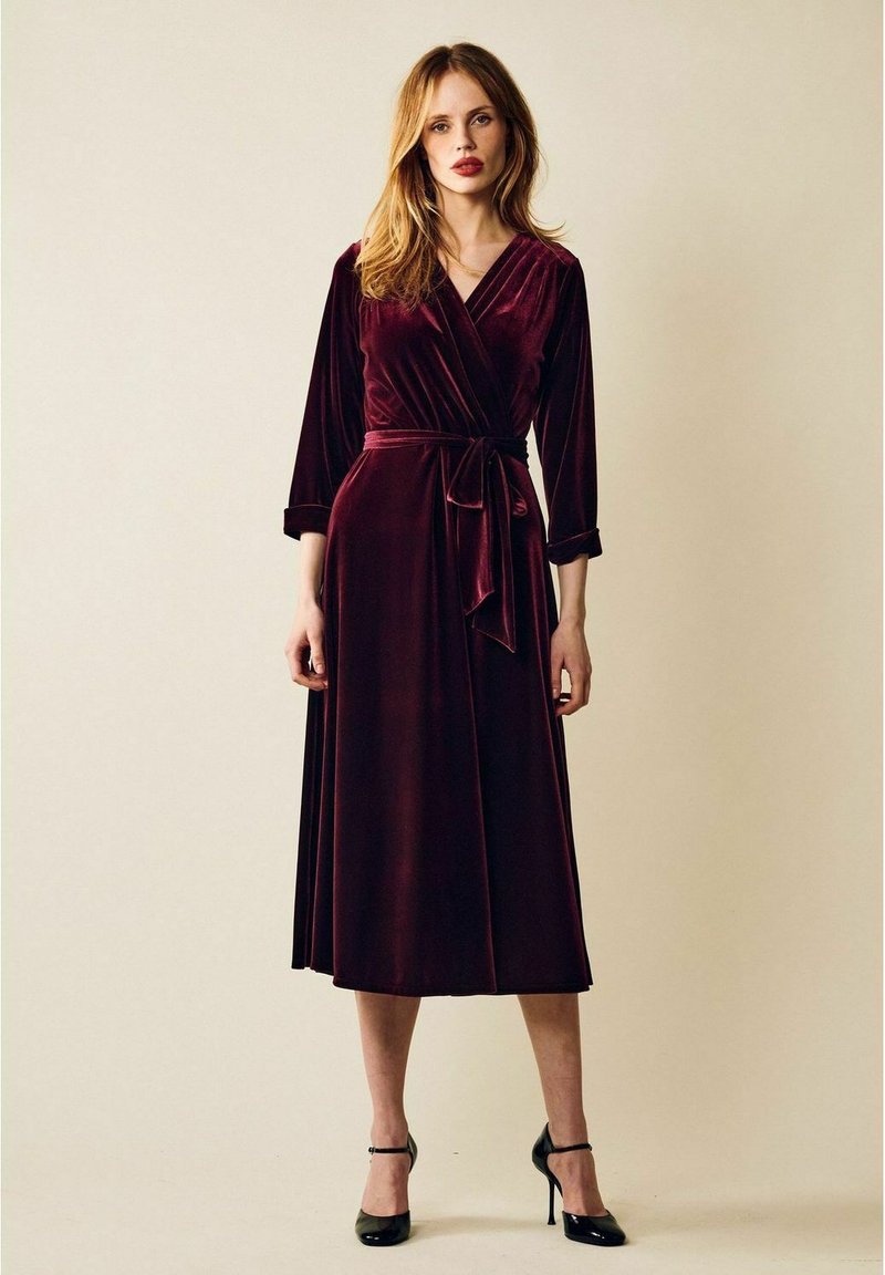 Burgundin samettinen kietaisumekko pitkillä hihoilla, solmittava vyötärö ja laskeutuva helma, joka ulottuu polven alapuolelle. Sileä pinta ja elegantti muotoilu.