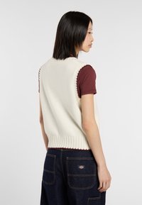 Chaleco de punto crema con dobladillo acanalado y bordes con forma de escarpa. Combinado con una camisa marrón, llevada sobre pantalones de mezclilla oscuros con bolsillos visibles.
